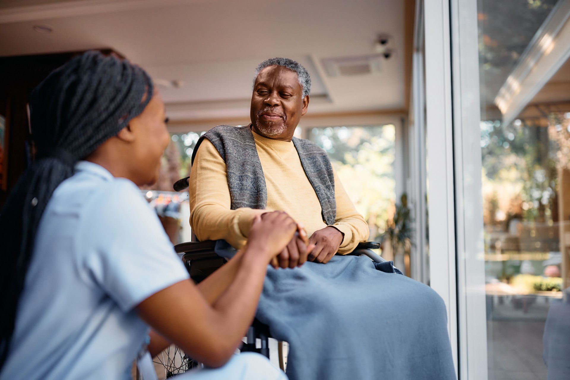 Smiling black senior man in wheelchair talking to a nurse while holding her hand in nursing home.
