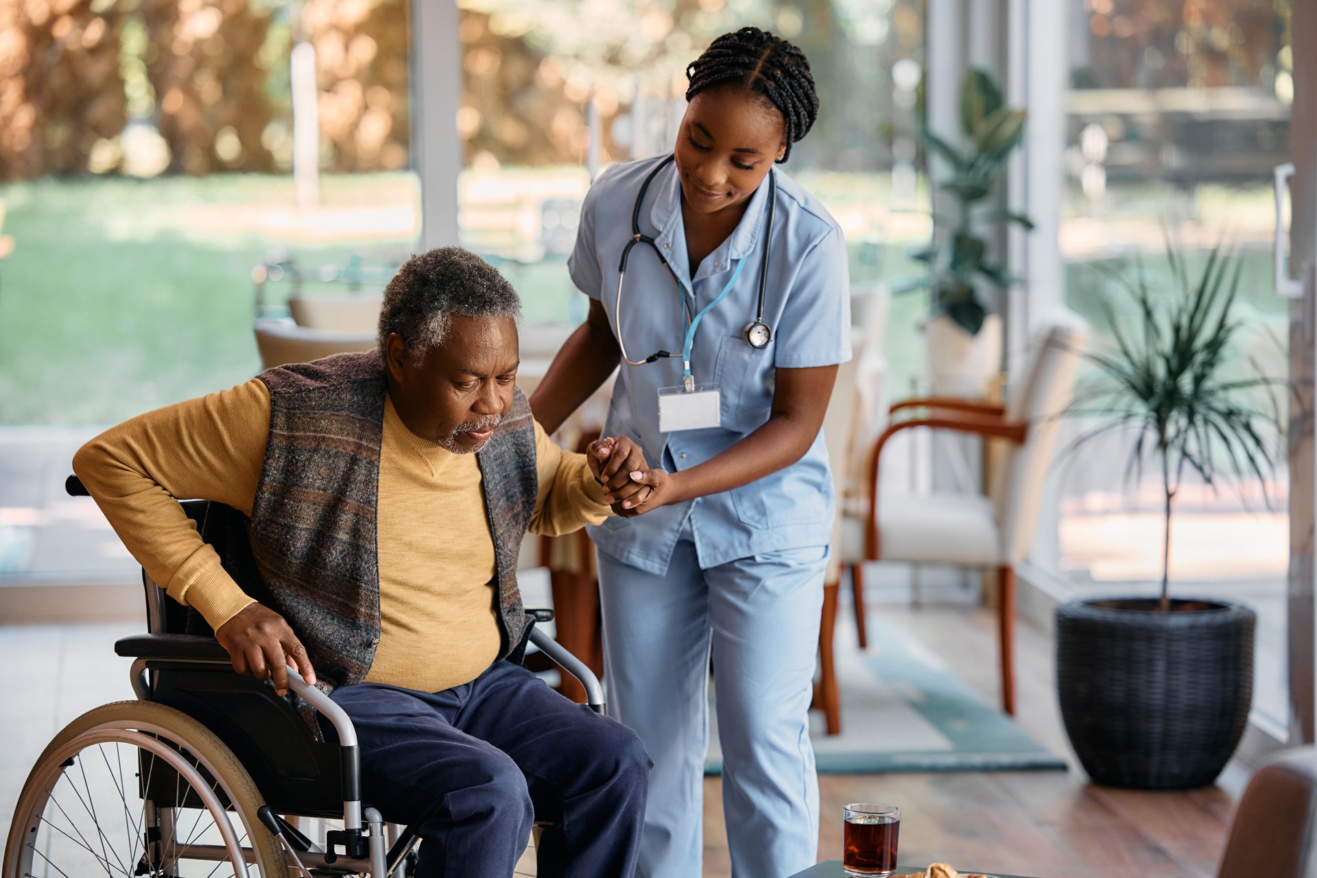 Young black nurse assisting senor man to get up from wheelchair at nursing home.